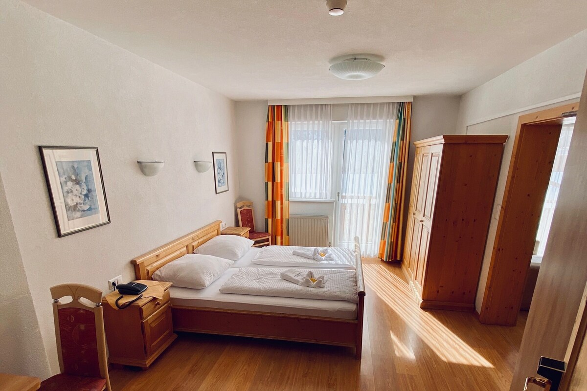 A hotel room with a wooden floor, a bed, a chair, a bedside table, a radiator, and a door.