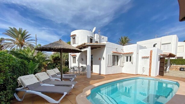 Zona junto a la piscina con sillas de descanso y una piscina, rodeada de edificios blancos y palmeras.