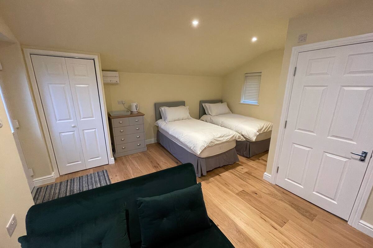 A compact bedroom with two single beds, a dresser, a green sofa, and a window with blinds.
