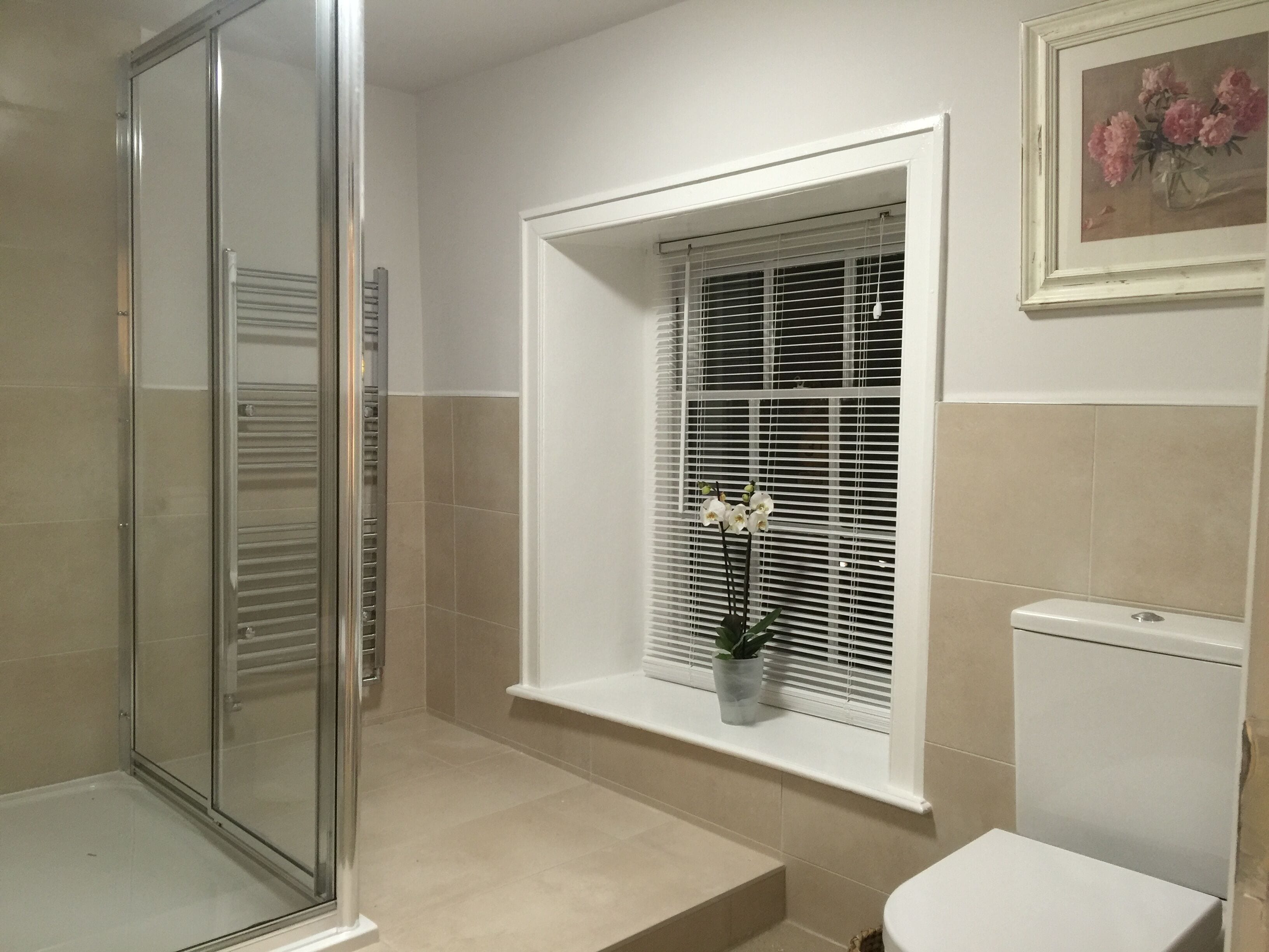 Une salle de bain avec une douche à parois de verre, une fenêtre avec des stores blancs et des toilettes.