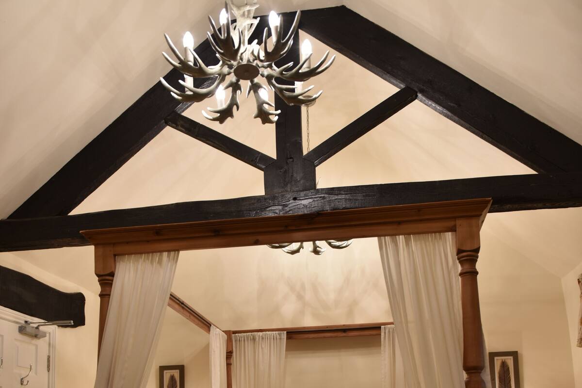 A four-poster canopy bed with a wooden frame and white drapes, a chandelier, and a plaid carpet.