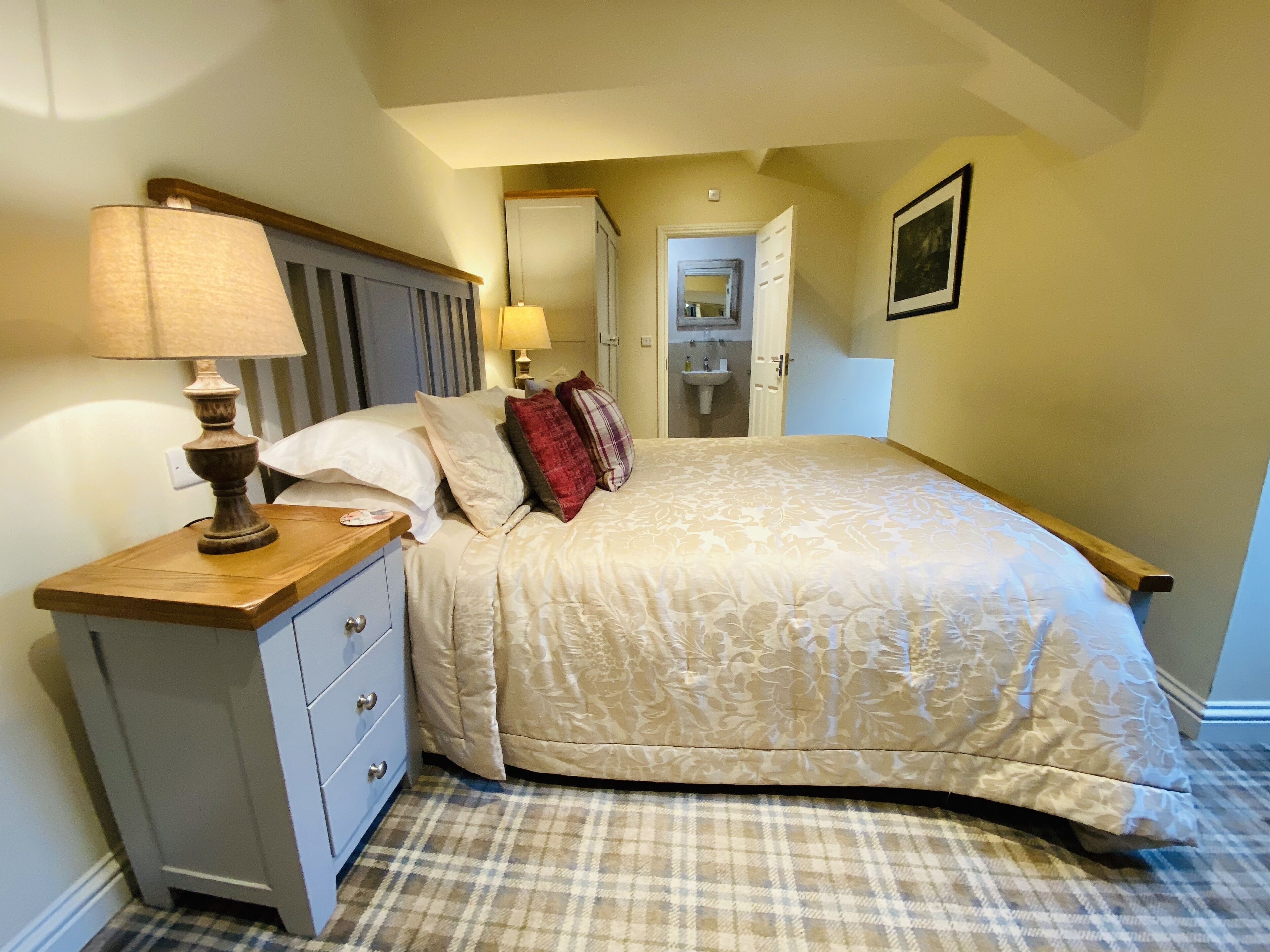 A bedroom with a bed, a nightstand with a lamp, a dresser, a bathroom visible through an open door, and a framed picture on the wall.