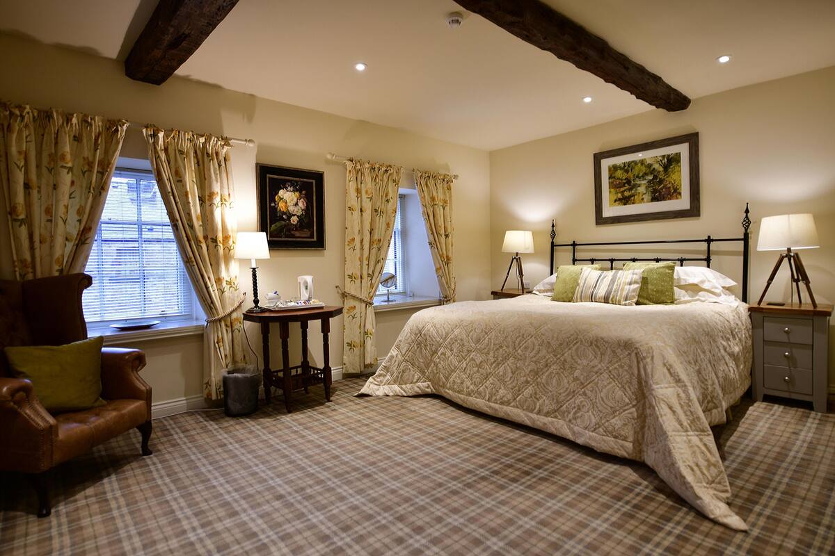 A bedroom with a large bed, two bedside tables with lamps, a chair, a small table with a lamp, and a window with curtains.