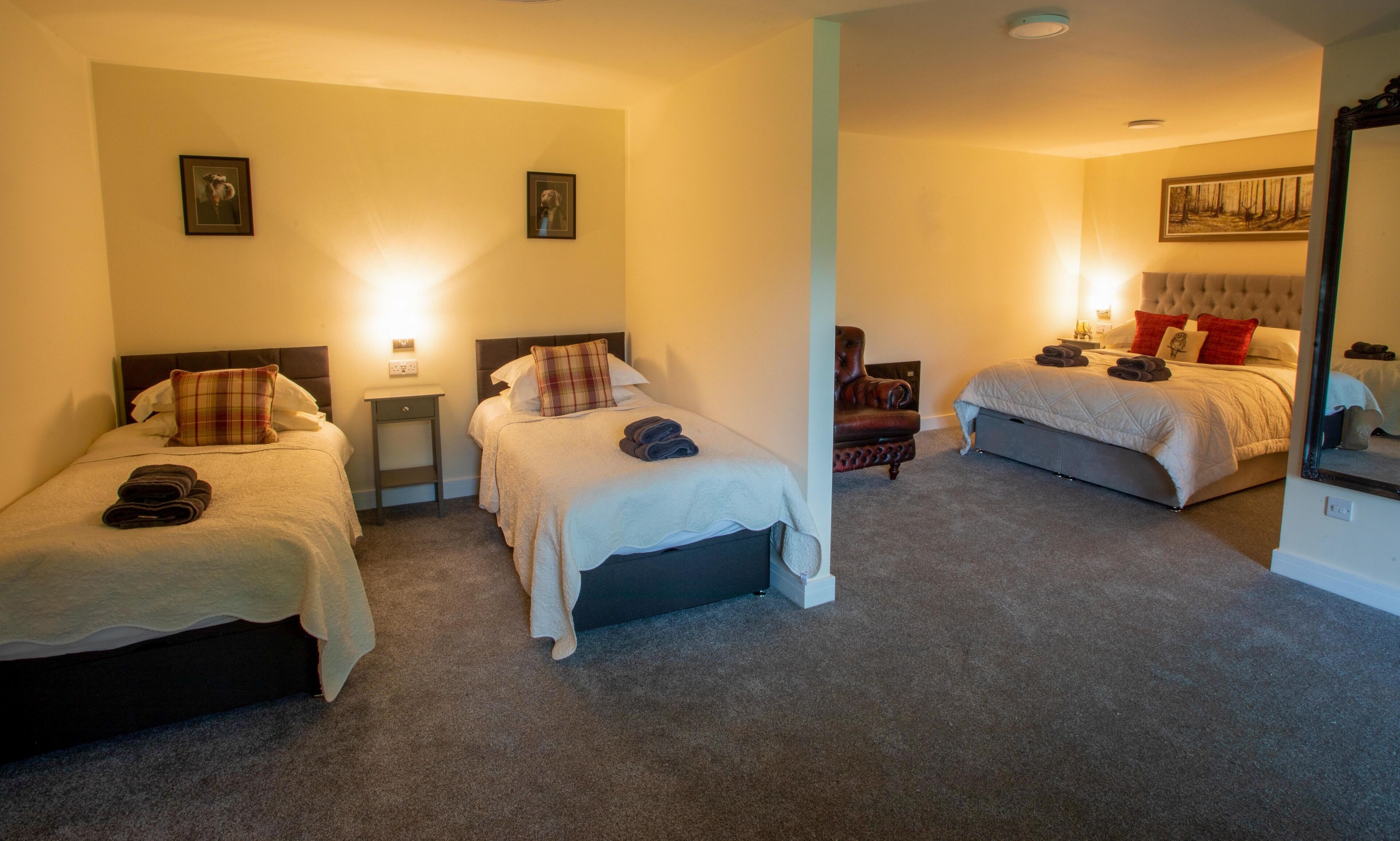 A hotel room with two single beds, a chair, and a larger bed. There are wall-mounted lights and a painting above the larger bed.
