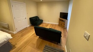 Een kamer met een houten vloer, een groene bank, een televisie op een houten dressoir en een witte deur.