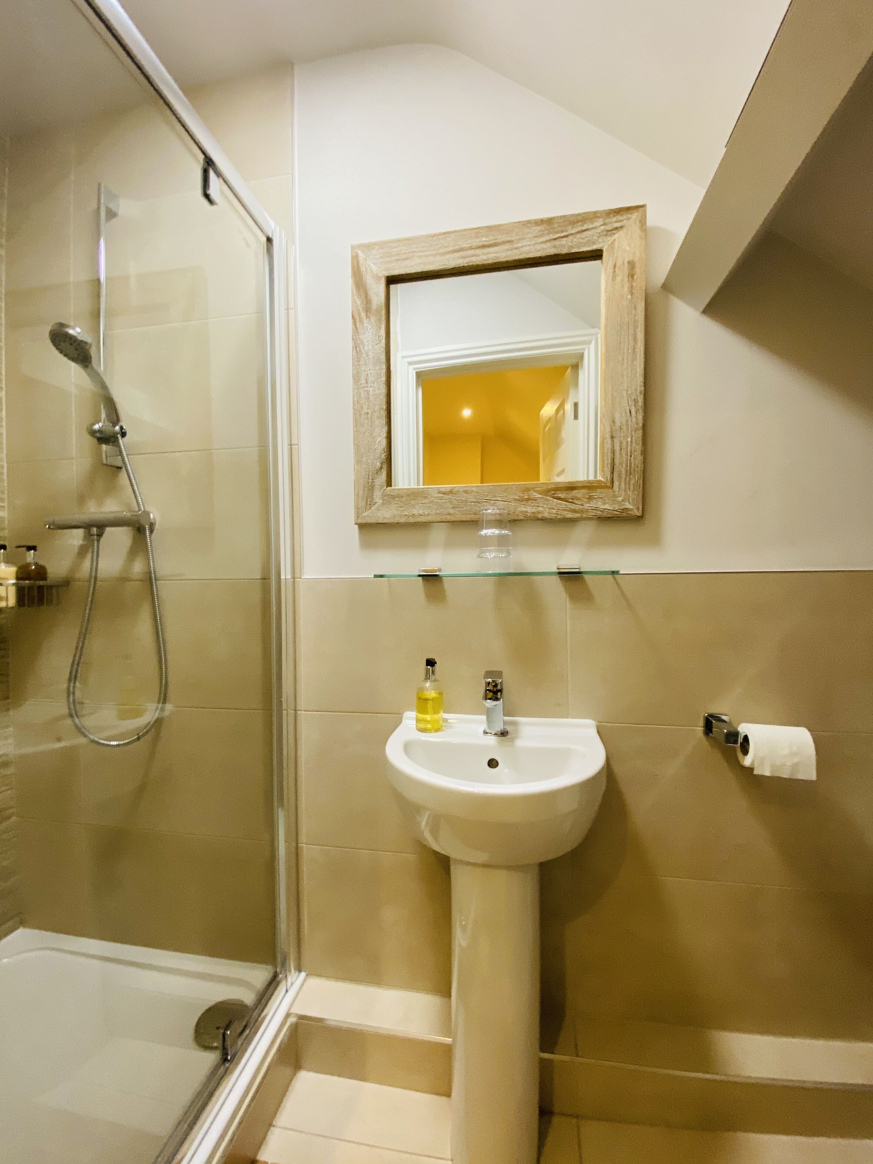 Un bagno con cabina doccia in vetro, lavabo su piedistallo, uno specchio con cornice in legno e porta carta igienica.