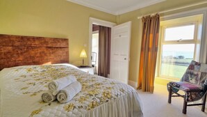 Habitación de hotel con una cama grande, una silla, vista al mar y una puerta que da a otra habitación.