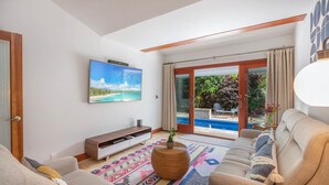 A living room with a TV, sofa, and a view of a pool and garden. - Makana Lei: Recently Upgraded Home w/ Private Pool, AC & Garden Views (Princeville)