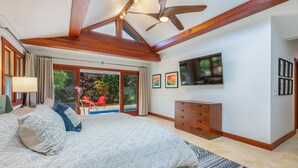 A bedroom with a large bed, a ceiling fan, a TV, and a view of a pool and garden through a sliding door. - Makana Lei: Recently Upgraded Home w/ Private Pool, AC & Garden Views (Princeville)