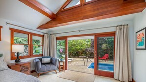 A spacious bedroom with a large bed, a sitting area with a chair and a table, a sliding glass door leading to a pool area, and a framed artwork on the wall. - Makana Lei: Recently Upgraded Home w/ Private Pool, AC & Garden Views (Princeville)