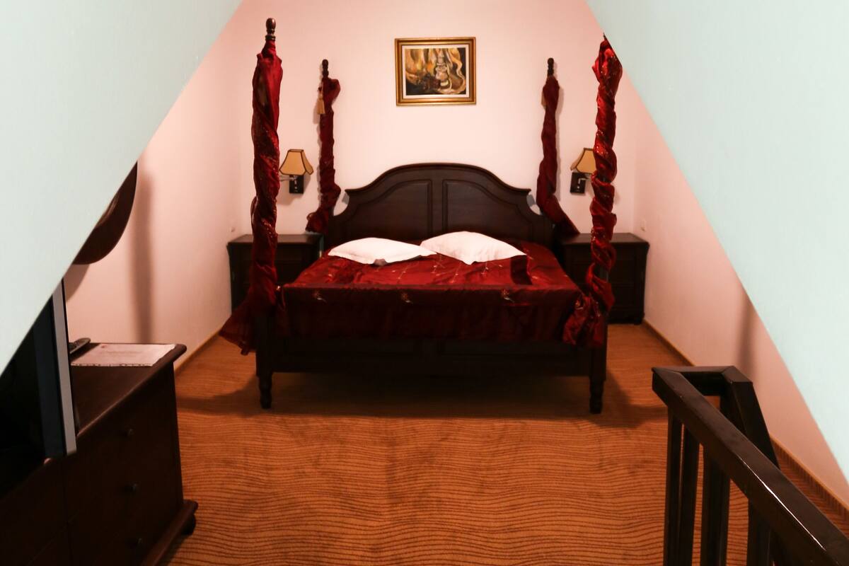 A bedroom with a four-poster bed, a wooden headboard, a red canopy, a nightstand with a lamp, and a framed picture on the wall.