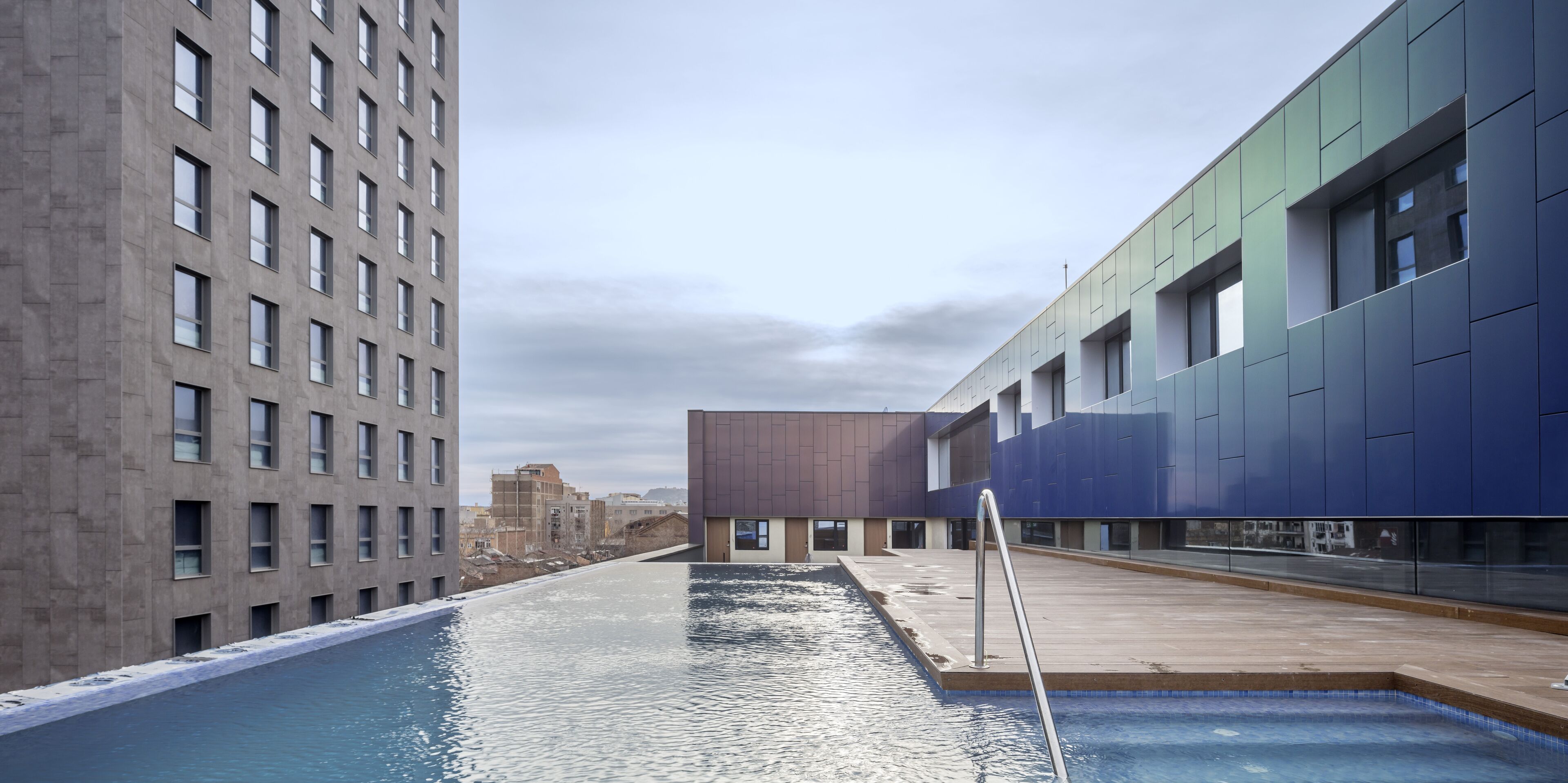 Un bâtiment moderne doté d’un espace piscine, comprenant une longue piscine avec un escalier en métal et offrant une vue sur le paysage urbain en arrière-plan.