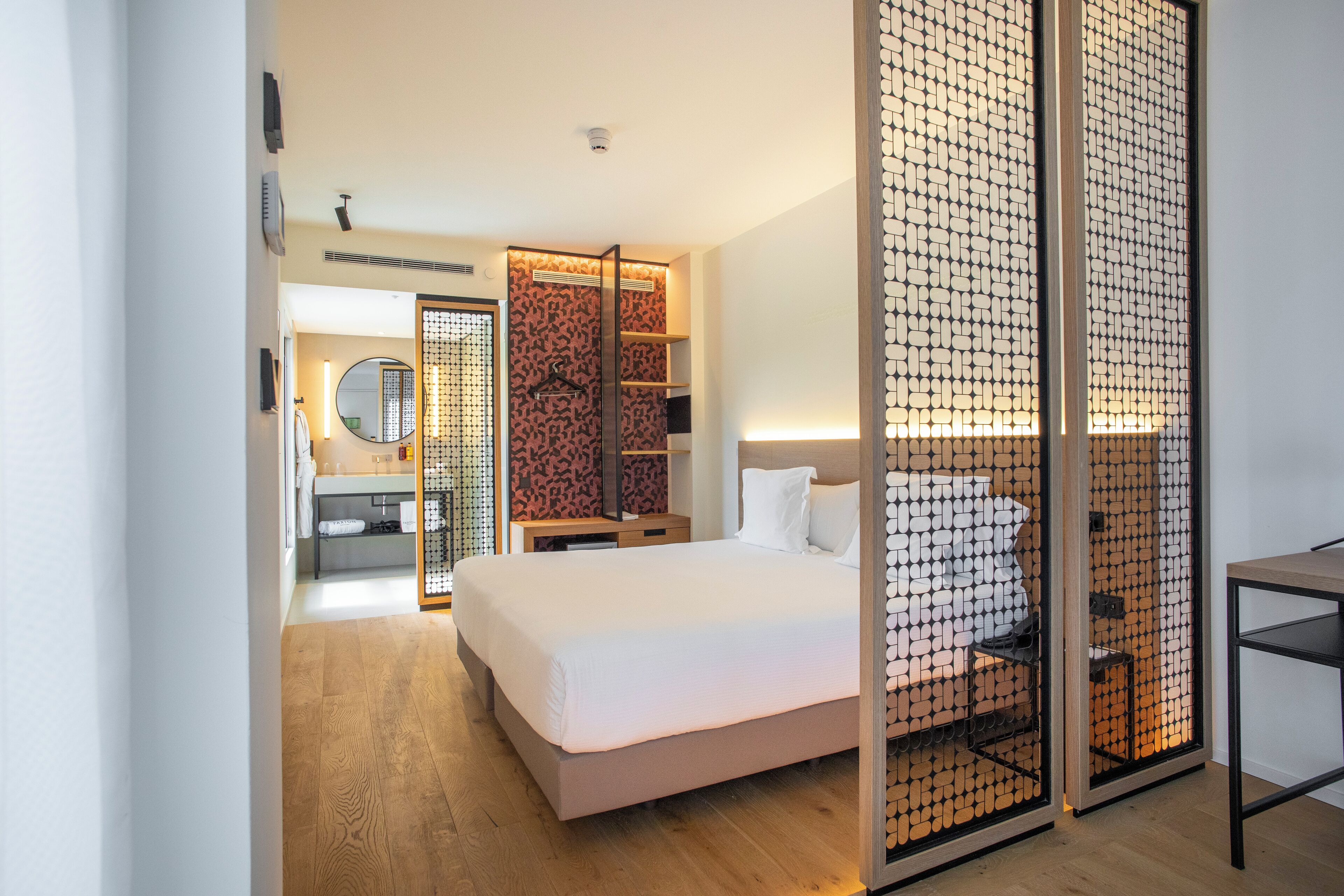 A modern hotel room with a large bed, a wooden floor, a bathroom with a mirror and sink, and a sliding door with a patterned design.