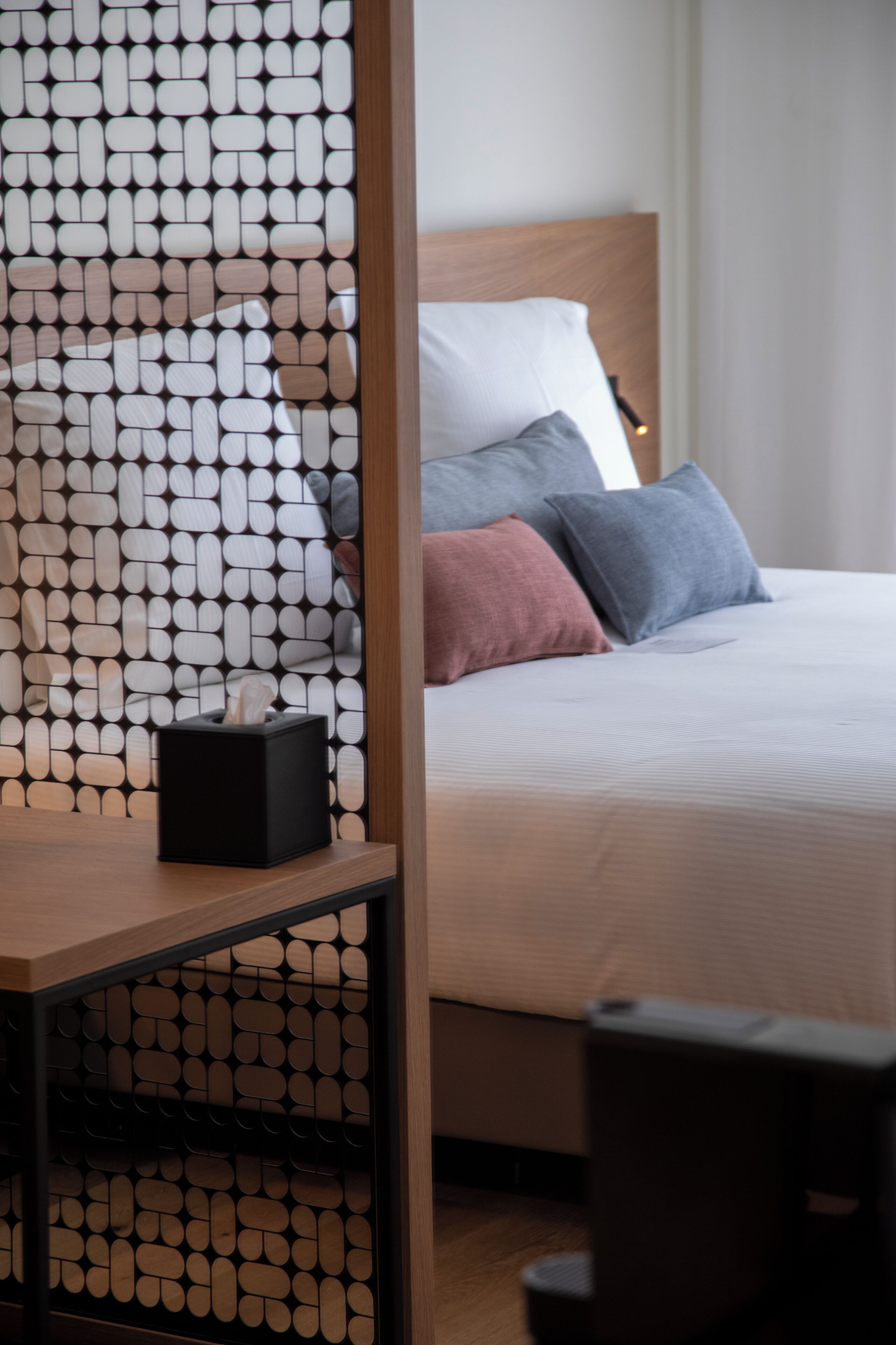 Una cama bien hecha con cabecera de madera, una mesita de noche con un dispensador de pañuelos de papel y un separador de ambientes con diseño.