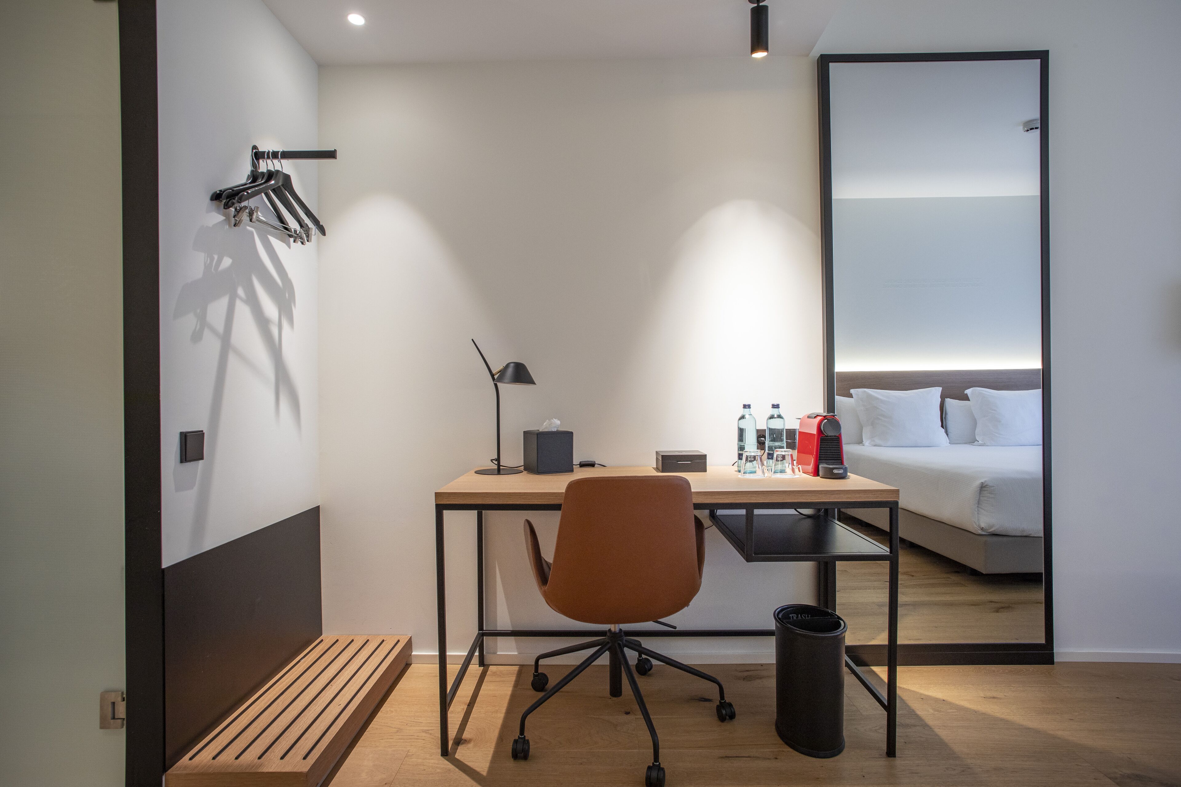 Una habitación de hotel moderna con un escritorio de madera, una silla de oficina individual, una cama con ropa blanca y un espejo grande en la pared.