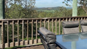 Een houten terras met een glazen tafel en twee rieten stoelen, met uitzicht op een pittoresk landschap met bomen en heuvels.