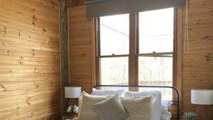 Een slaapkamer in een houten hutje met een metalen bedframe, witte beddengoed en een raam met uitzicht op groen.