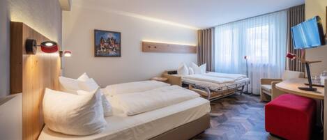 A hotel room with two beds, a desk with a chair, a TV, and a red ottoman.