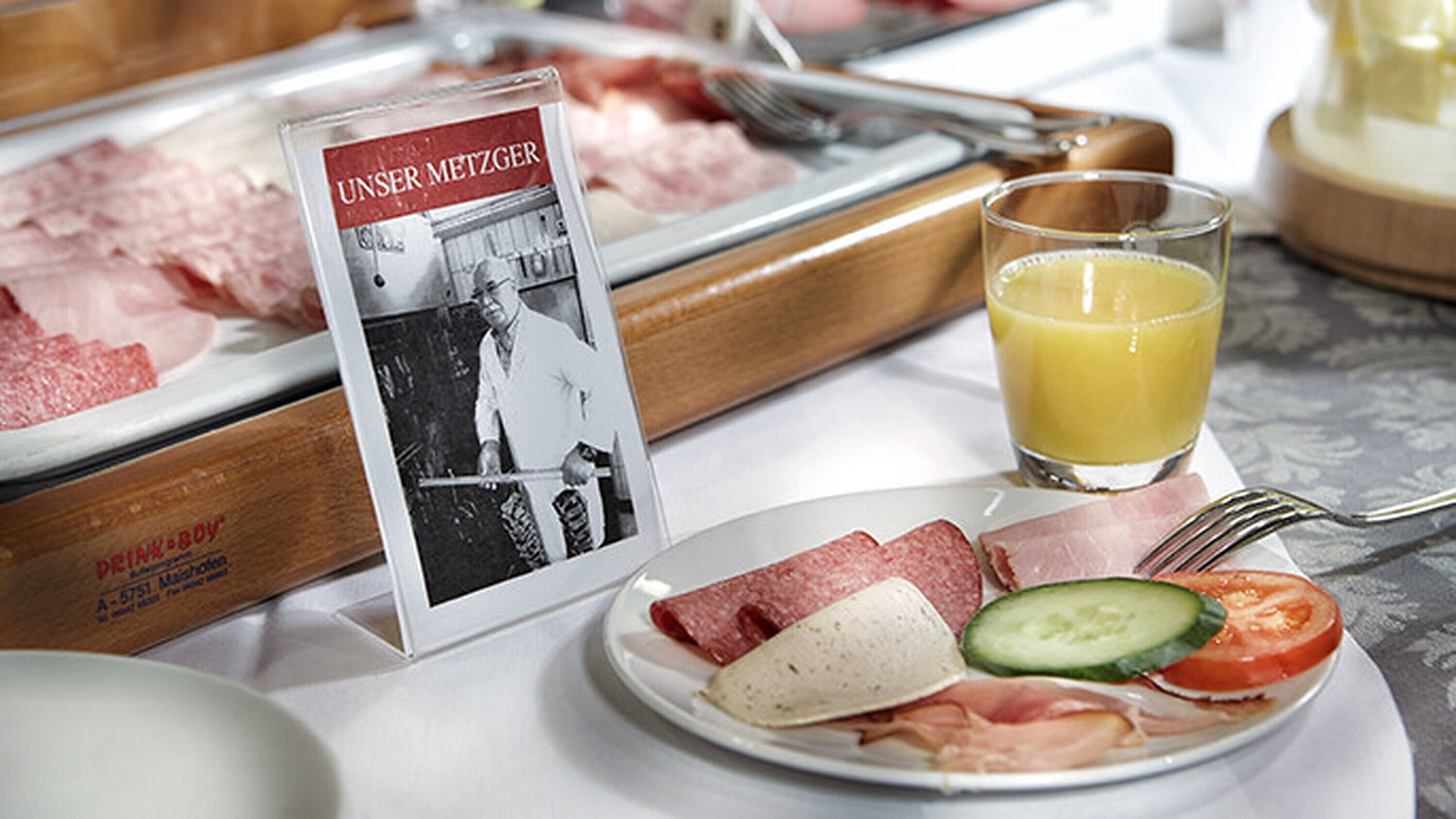 A breakfast setting with a plate of meats, cheese, and vegetables, a glass of orange juice, and a sign with a photo.