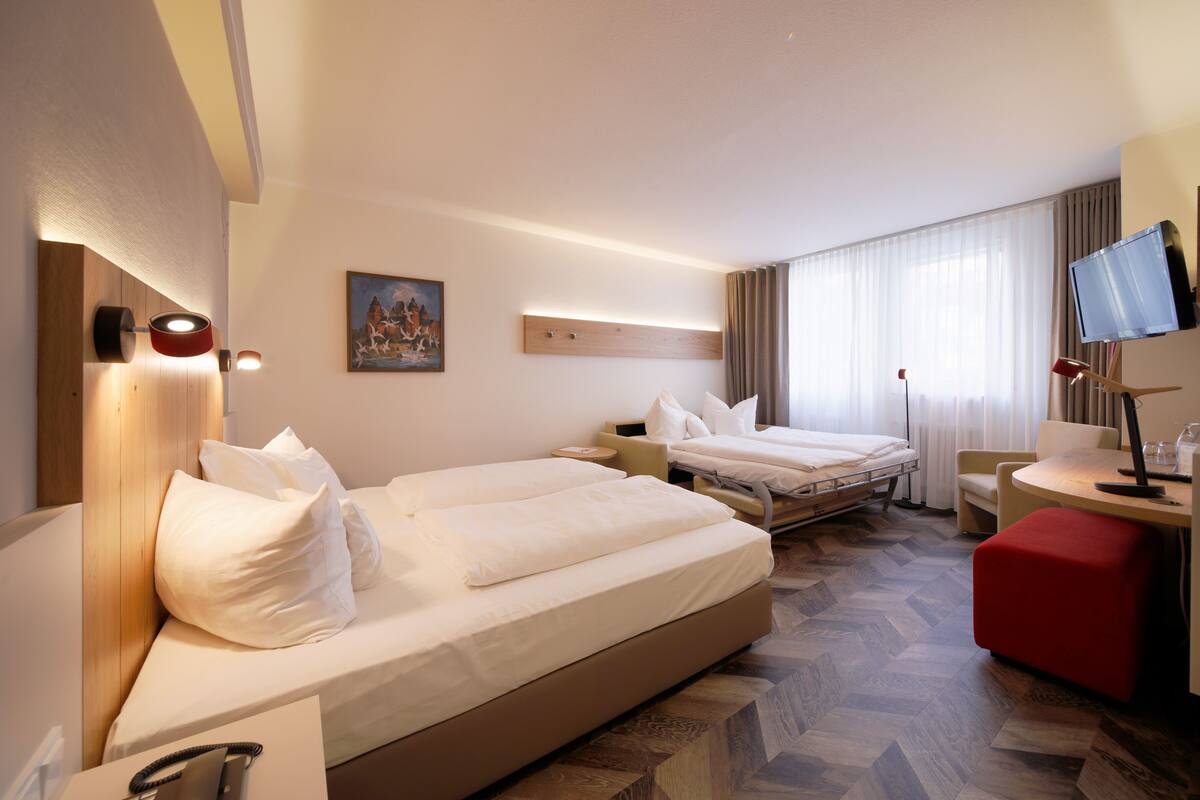 Habitació d'hotel amb un llit gran, un llit més petit, una taula de treball, una cadira, un televisor i un quadre penjat a la paret.