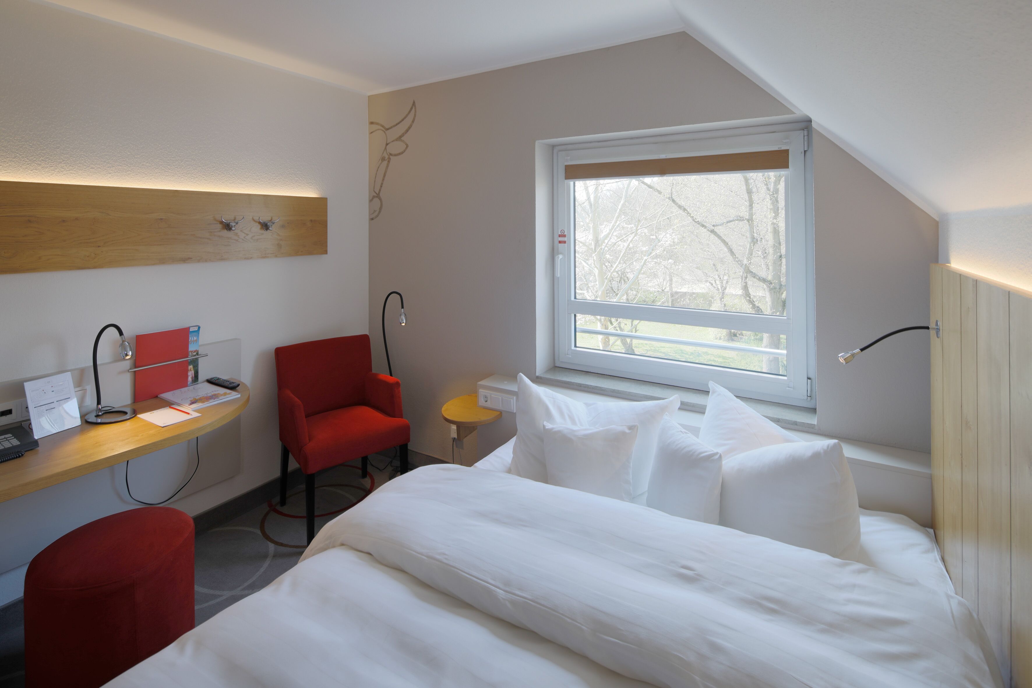 Habitación de hotel moderna con cama, escritorio con ordenador, una silla roja y ventana con vista a árboles.