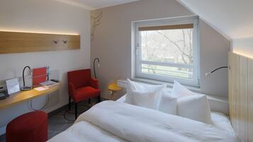 Habitación de hotel moderna con cama, escritorio con ordenador, una silla roja y ventana con vista a árboles.