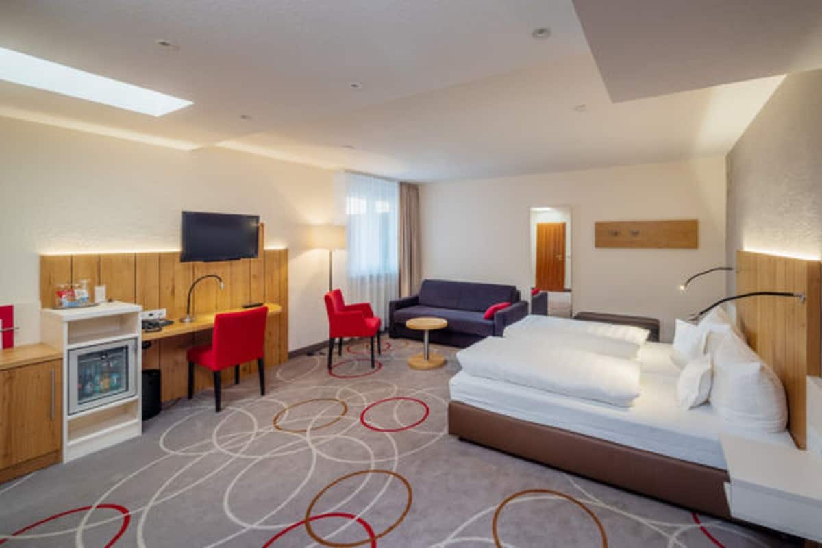 Habitació d'hotel amb un llit, una taula de treball amb una cadira, un sofà, un televisor i un minibar.