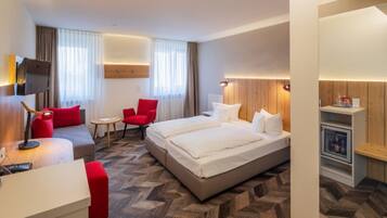 A hotel room with a bed, two red chairs, a small table, a TV, and a minibar.