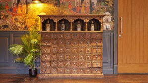 A wooden chest of drawers with multiple compartments, a potted plant, and a mural in the background. - The Strutt Arms (Belper)