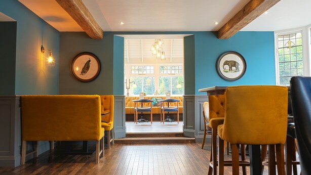 A restaurant with a blue wall, wooden beams, and yellow chairs. There are framed pictures on the wall and a chandelier above the seating area. - The Strutt Arms (Belper)
