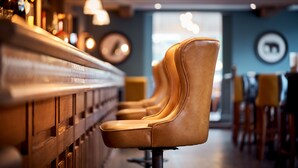 A row of leather chairs with a high backrest and a metal base, positioned in a bar area with a wooden counter and a blue wall with circular wall art. - The Strutt Arms (Belper)