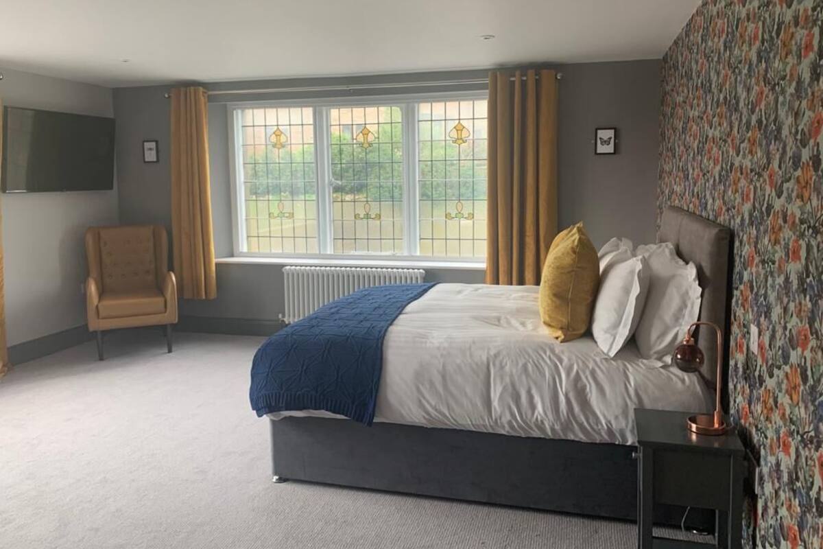A bedroom with a bed, a chair, a radiator, a window with curtains, and a wall with a floral pattern.