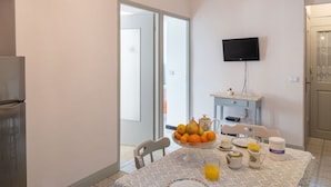 Un coin repas avec une table mise pour le petit-déjeuner, un saladier de fruits, une cafetière et un téléviseur fixé au mur.