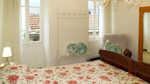 Une chambre avec une couvre-lit à motifs floraux, un étendoir à linge en métal blanc, une chaise en bois, un lustre et une fenêtre donnant sur un toit recouvert de tuiles.