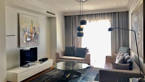 A modern living room with a flat-screen TV on a white console, a round glass coffee table, a beige sofa with grey cushions, and a floor lamp.