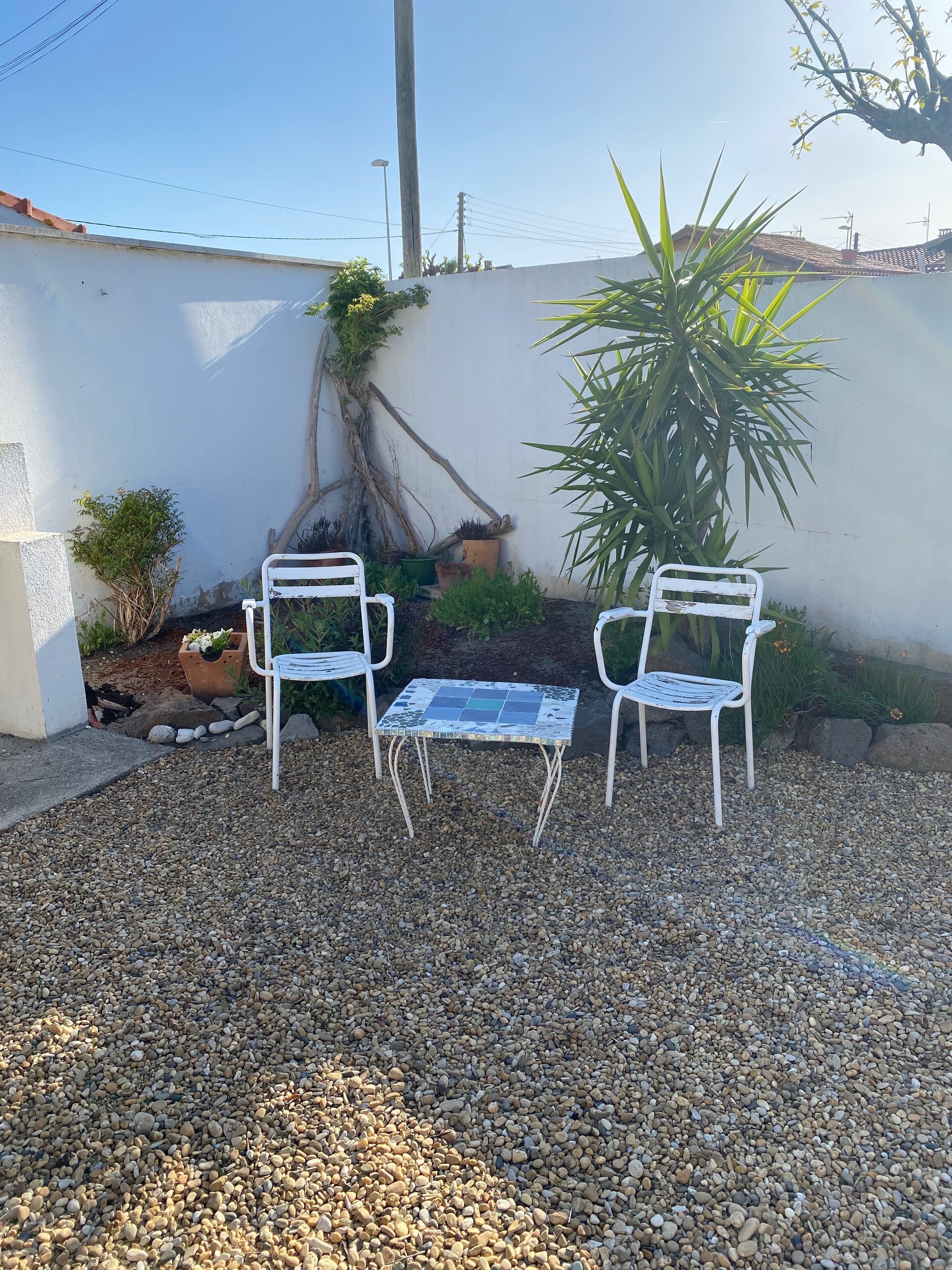Een patio met twee witte metalen stoelen en een kleine tafel, omringd door kleine steentjes en potplanten.