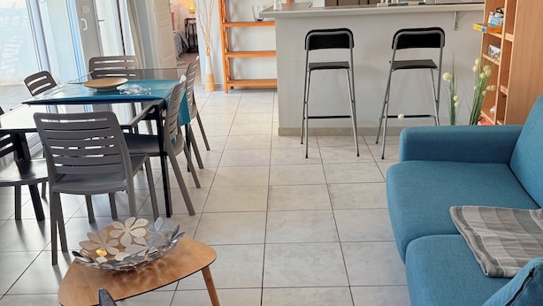 A modern living room with a blue sofa, a dining area with a table and chairs, a kitchen area with a bar, and a large window with curtains. - Air-conditioned villa with garden in the center of Marseillan (Marseillan)