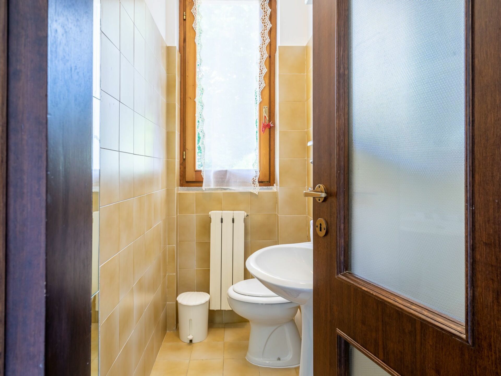 Un baño con inodoro, un pequeño cubo de basura y una ventana con cortinas de encaje.