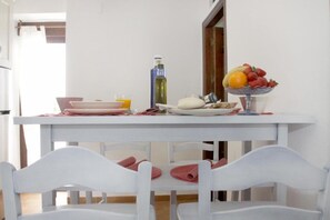 A dining area with a white table set for two, a bowl of fruit, a bottle, and plates with a glass of orange juice.