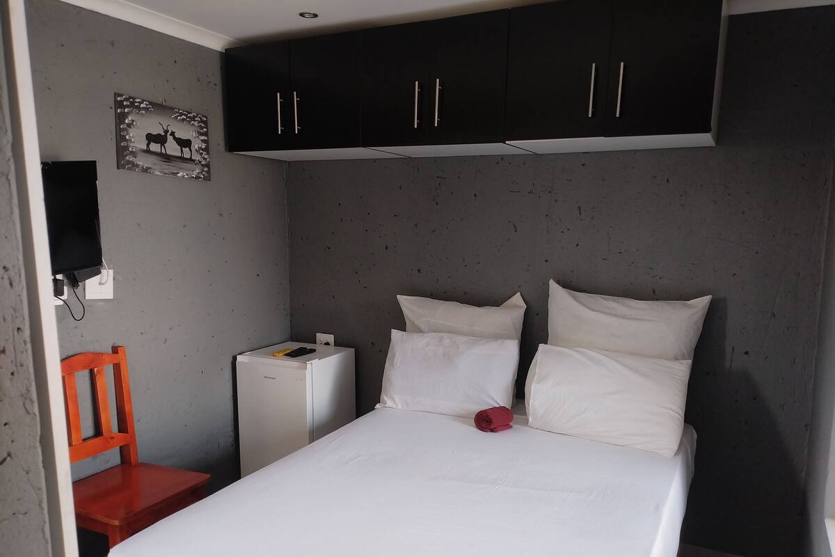 A bedroom with a bed, white pillows, a red chair, a small white bedside table, and a black cabinet above the bed.
