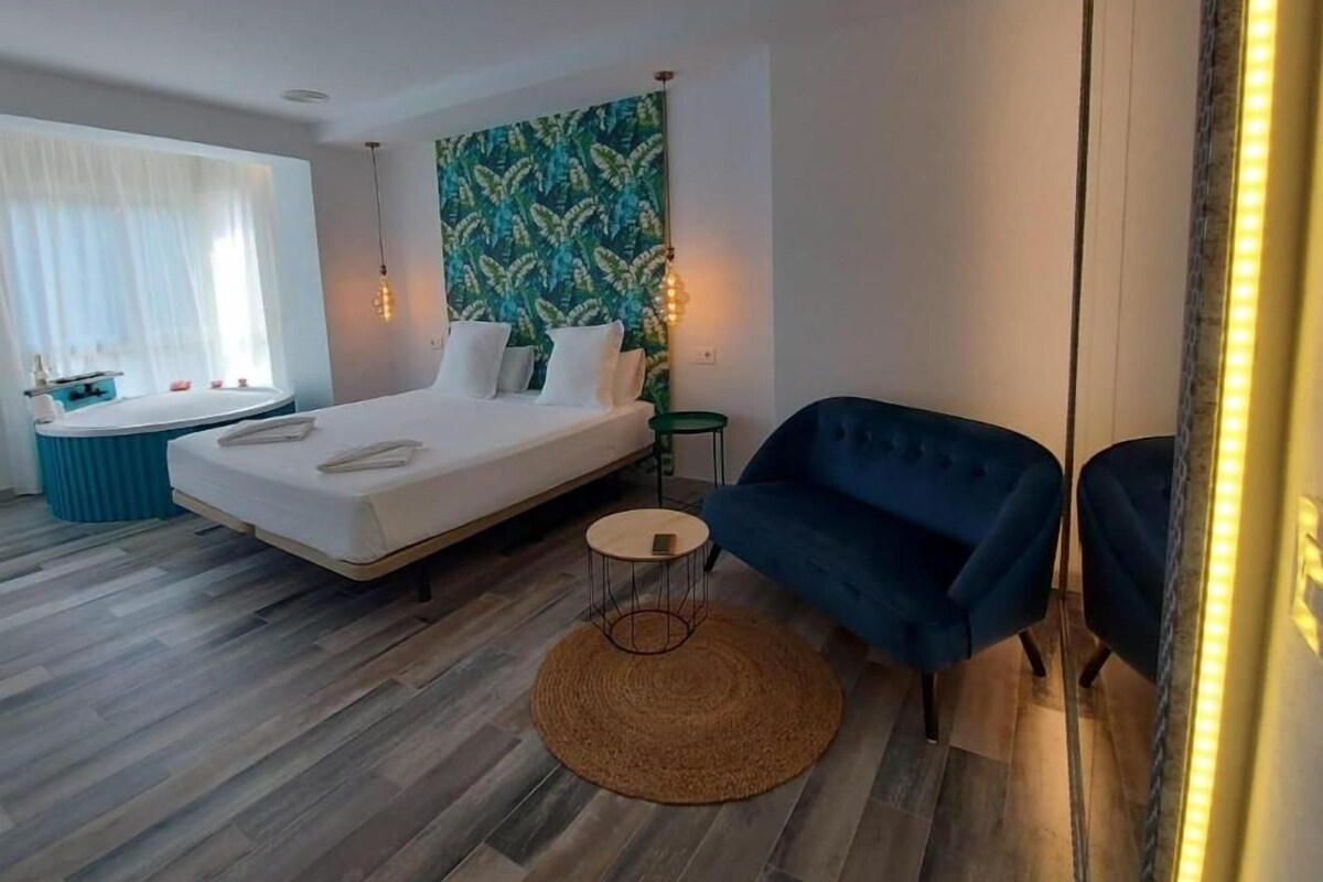 A modern bedroom with a bed, a blue sofa, a small round table, and a wall with a leafy pattern.