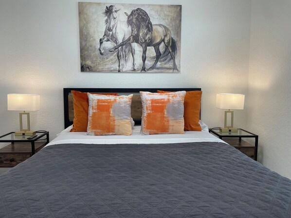 A neatly made bed with a quilted grey blanket, two orange and white pillows, and a painting of horses on the wall above.