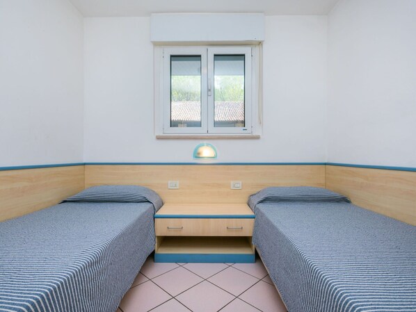 A small room with two single beds, a window, a desk with a lamp, and a tiled floor.