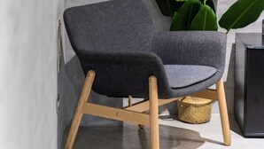 Una silla moderna con patas de madera y asiento de tela oscura, ubicada frente a una pared de concreto con una planta verde grande cerca.