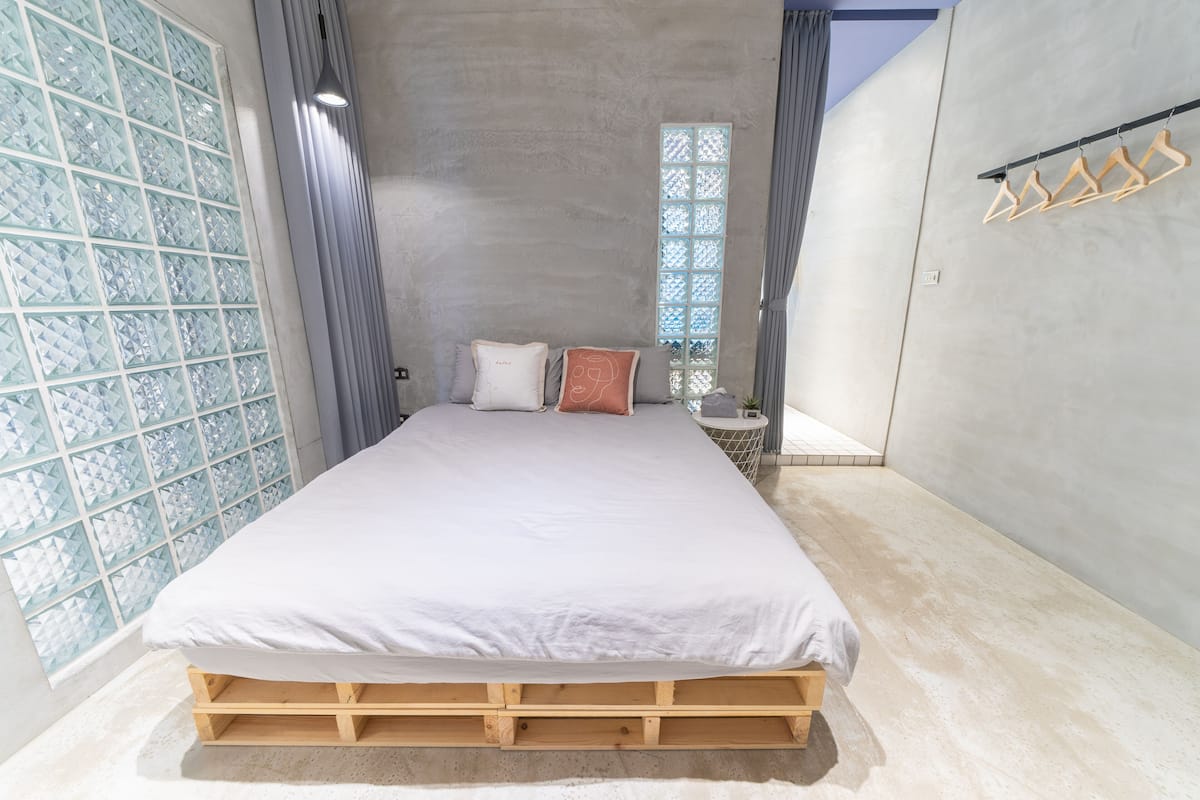 Ein Schlafzimmer mit hölzernem Bettgestell, einem großen Fenster mit geometrischem Muster und einem hängenden Kleiderständer.