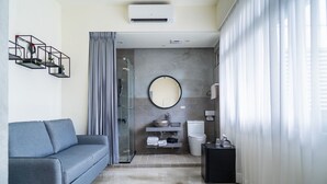 Una habitación de hotel moderna con un sofá gris, un espejo redondo, un baño con inodoro y lavamanos, y un box de ducha de vidrio.