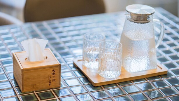 Una caja de pañuelos de madera, dos vasos y una jarra de vidrio sobre una mesa con azulejos.