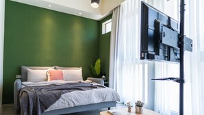 Un dormitorio con una cama, un televisor montado en la pared, una ventana con cortinas y una mesita de noche con una planta.