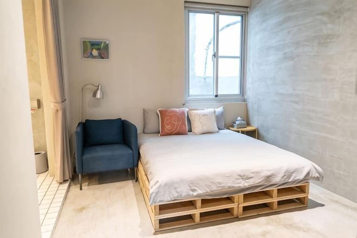 A bedroom with a bed, a chair, a bedside table, a lamp, and a window.