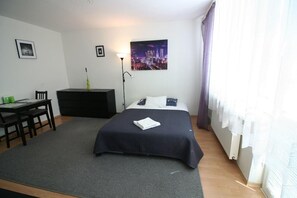 Un dormitorio con cama, escritorio, silla, cómoda y un cuadro de un paisaje urbano en la pared.